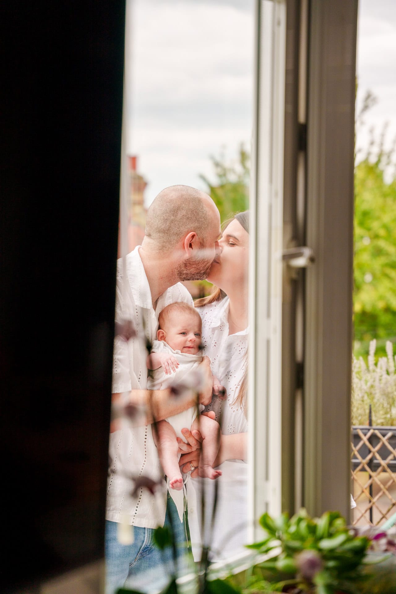 Smiling family sharing a moment of love and affection at a bright, airy window, with lush greenery outside and a decorative plant in the foreground.