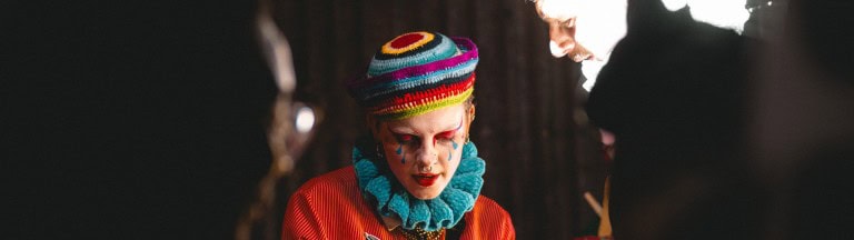 Brightly dressed individual with colourful makeup and accessories, engaging with others in a creative and artistic environment.