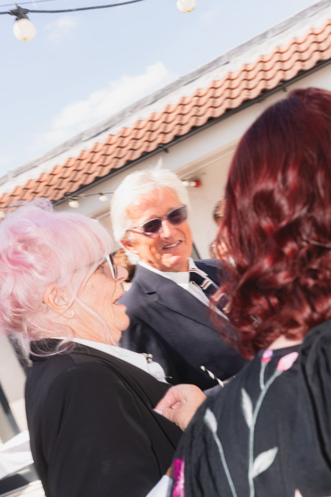 Colourful group of people smiling and holding hands during an outdoor social gathering with a charming building and string lights in the background.