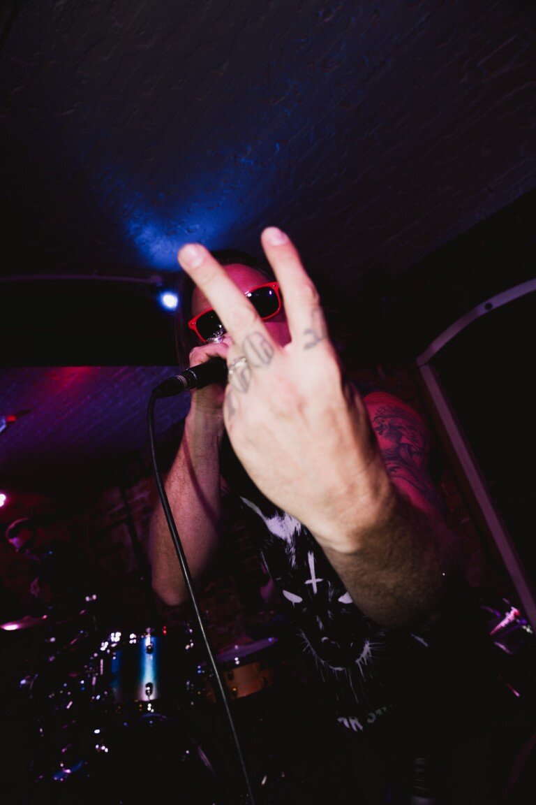 Vibrant performer in darkened venue stands close to the camera, wearing red sunglasses and holding a microphone, creating an energetic live music atmosphere.