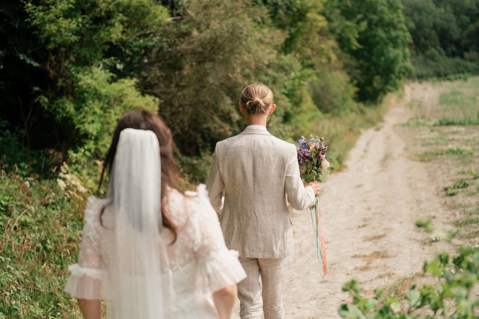 Elegant wedding photography of a bride and groom walking outdoors on a scenic dirt path surrounded by lush greenery, captured with natural light.