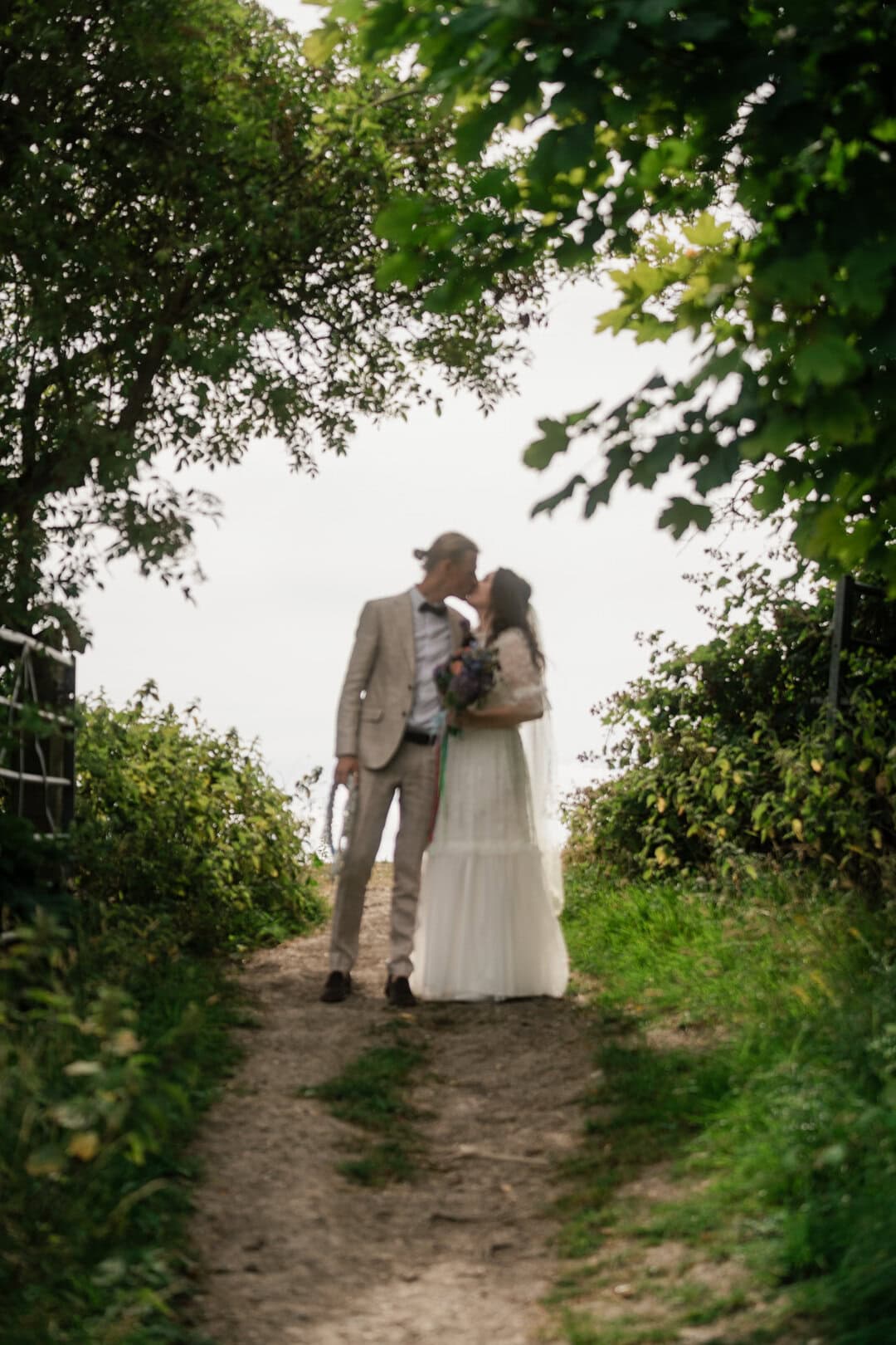 Romantic wedding couple sharing a kiss underneath lush green trees on a nature trail.