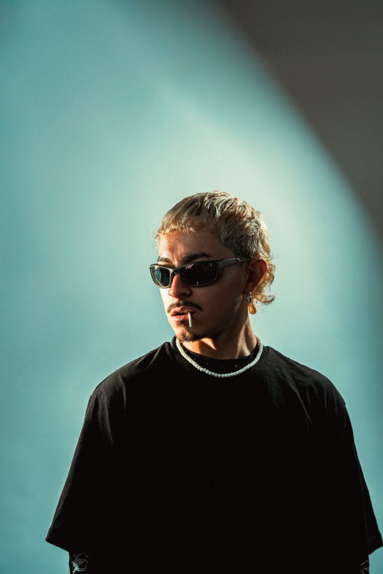Close-up portrait of a young man with blonde hair, wearing dark sunglasses, a black t-shirt, and a pearl necklace, with dramatic lighting and colourful background.