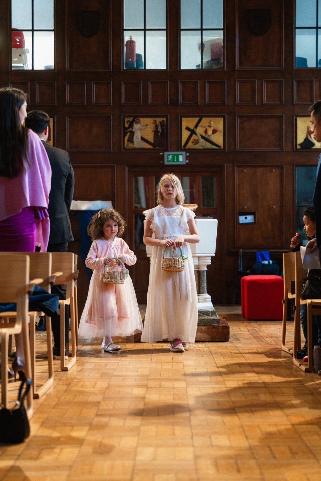 Delicate young girls in pastel dresses holding small baskets walk down the aisle of a church during a ceremonial event, surrounded by seated people.