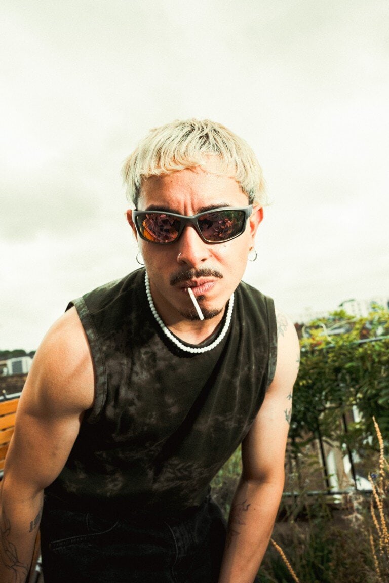 Close-up portrait of a punk man with bleached hair, dark sunglasses, smoking a cigarette outdoors against a cloudy sky.