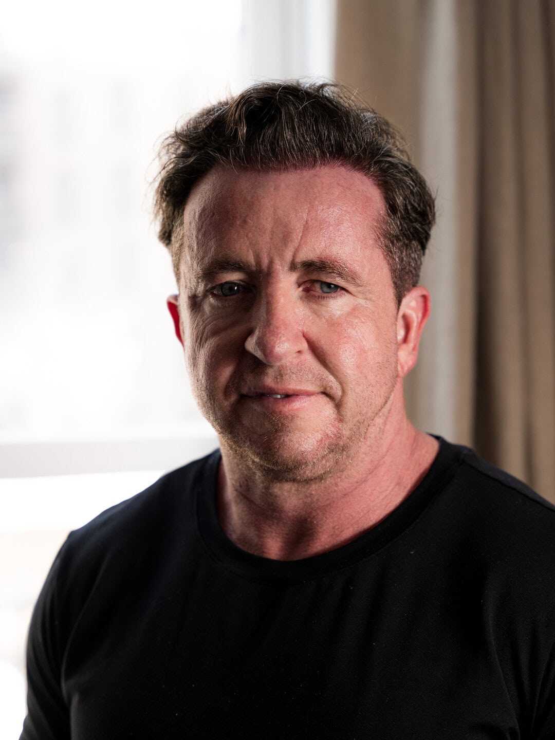 Close-up portrait of a middle-aged man with dark hair and blue eyes inside a modern apartment.