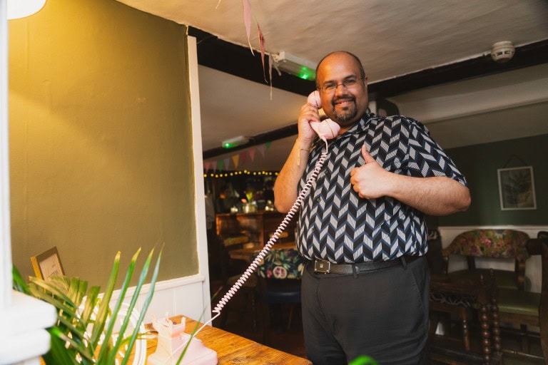 A person with a beard, glasses, and a patterned shirt giving a thumbs-up while using a vintage pink rotary phone at a social gathering.