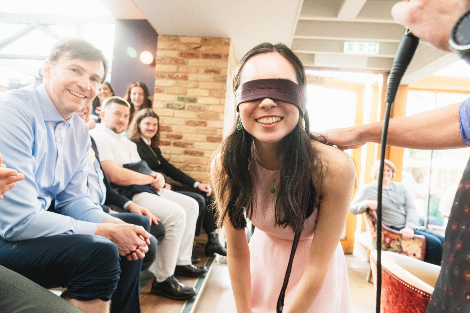 Blindfolded person smiling during a group activity at a social gathering in a cosy, light-filled space with exposed brick walls.