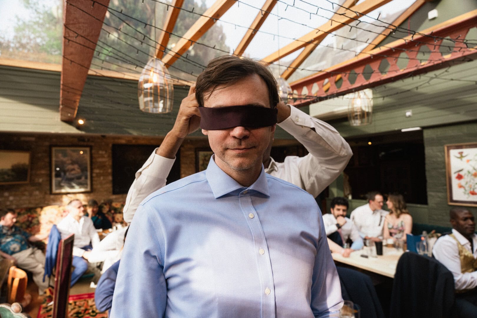 Blindfolded person at social gathering inside a rustic venue during daytime.