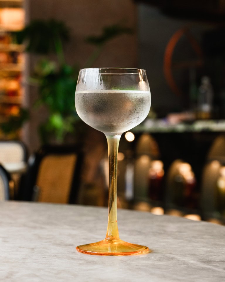 Shimmering cocktail glass filled with a clear beverage, condensation on the exterior, set against a cozy, dimly lit bar background.