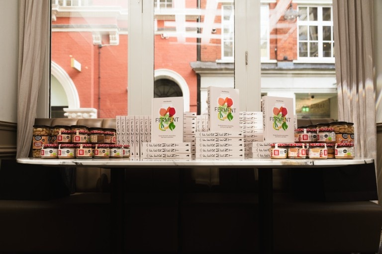 Sunny indoor display of fermented food jars and recipe books on a marble table in front of a large window showcasing a brick building.