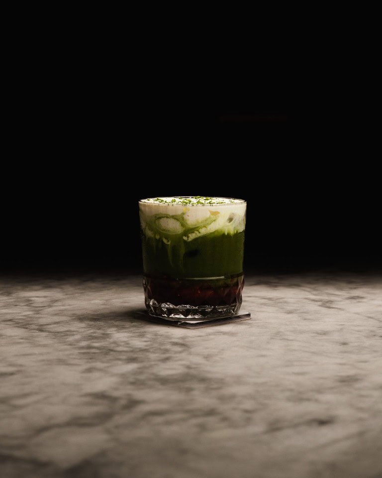 Butterscotch and matcha layered cocktail with whipped cream and green garnish in a clear, textured glass on a marble surface against a black background.