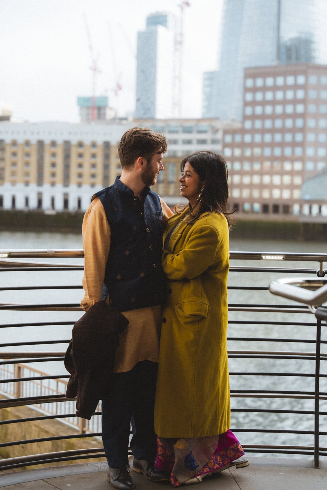 A couple of individuals smiling and sharing an intimate moment on a riverside promenade with a cityscape backdrop, including modern high-rise buildings and cranes.