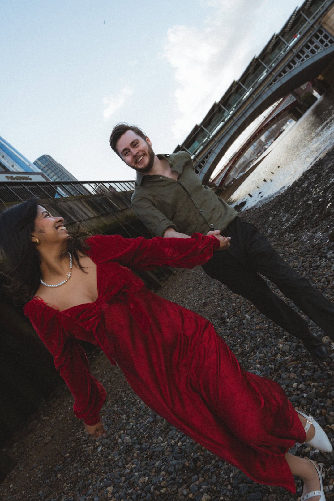 Vibrant red velvet dress worn by a smiling woman holding hands with a cheerful man beside a river under a bridge.