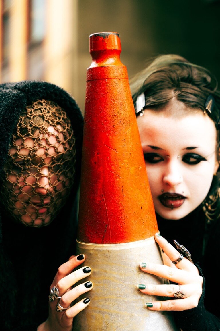 Bright orange traffic cone held by a person with dark hair and makeup, surrounded by alternative fashion accessories.
