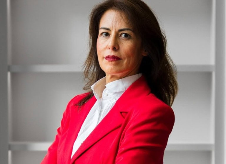 Confident professional woman with crossed arms in a red blazer and white shirt standing in a modern office space.
