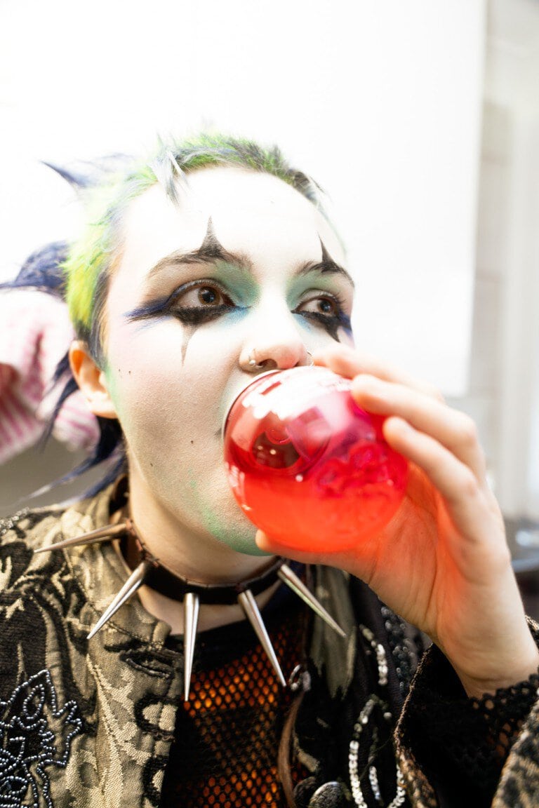 Vibrant individual with bold makeup and spiked jewelry enjoying a glass of red drink, showcasing alternative style and expressive fashion.
