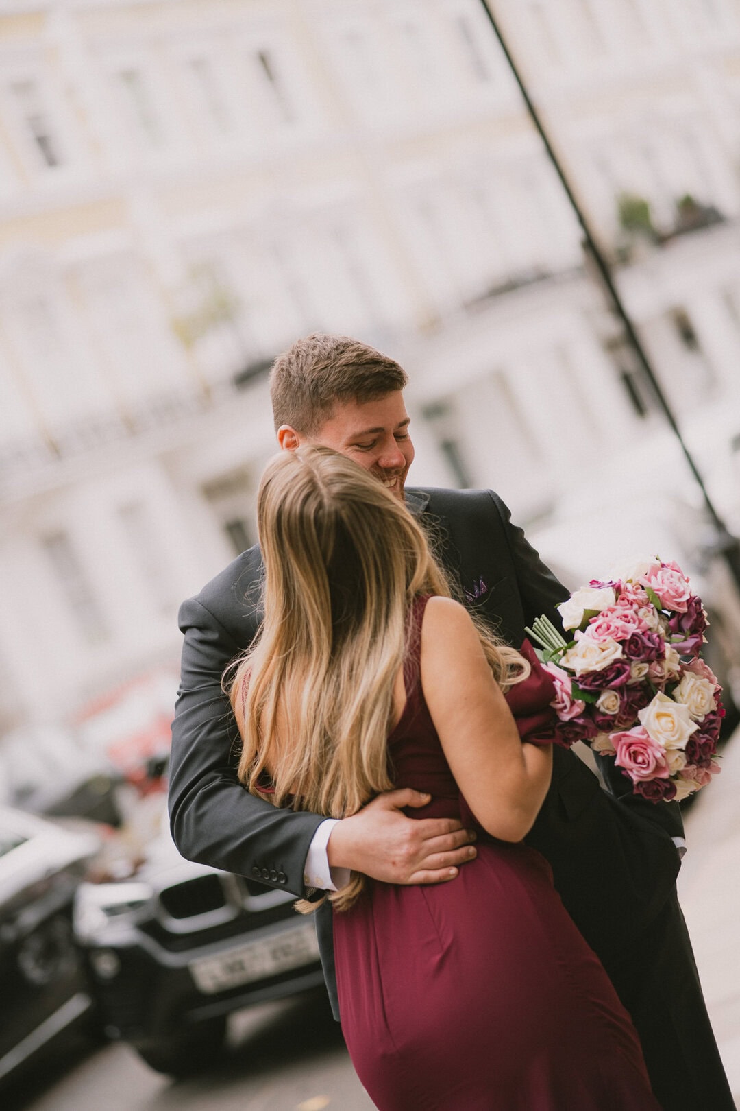 Smile, embrace, soft focus, urban background, romantic moment, formal attire, floral bouquet, happy couple, city street, wedding celebration, affectionate gesture, joyful expression, stylish clothing.