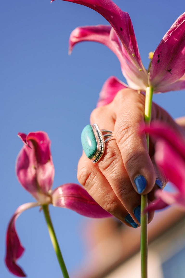 Vivid turquoise ring with silver accents worn on a person's finger, holding vibrant pink lilies against a clear blue sky background.