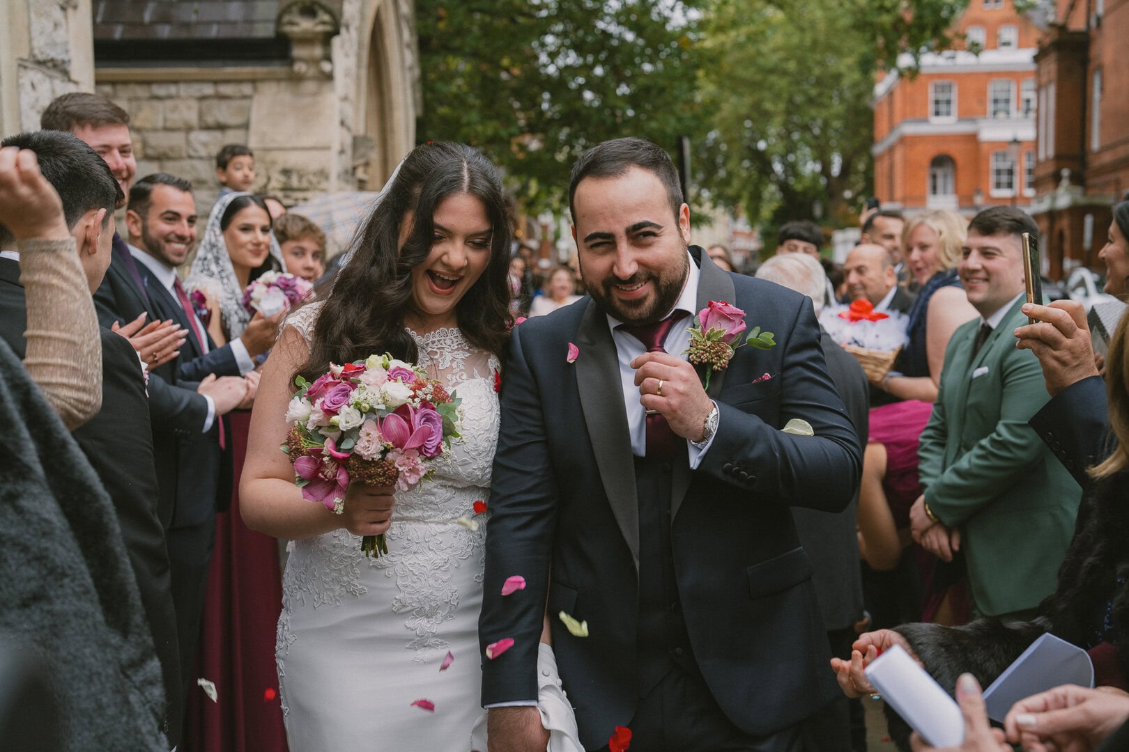 Bride and groom celebrating wedding outdoors with friends and family, colourful flowers, joyful atmosphere, historic building backdrop, autumn foliage.