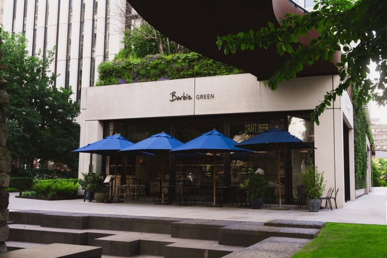 Bright modern café exterior with outdoor seating, blue umbrellas, lush greenery, and urban architecture in the background.