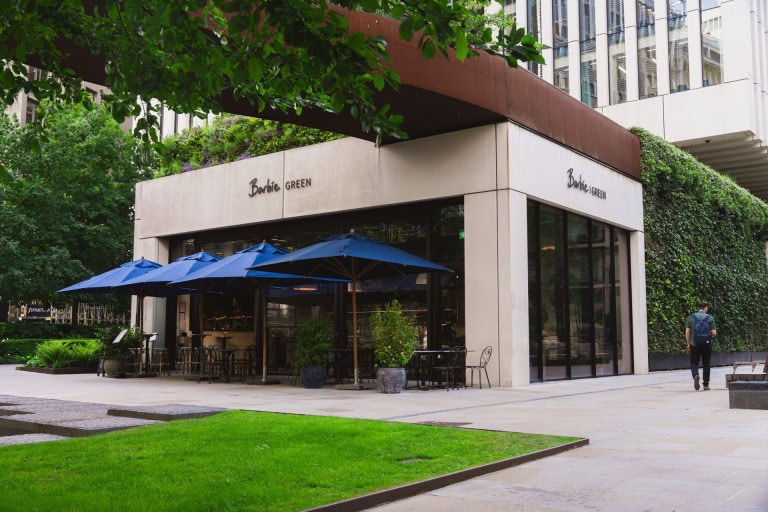 Modern shopping centre exterior featuring a café with blue umbrellas, lush greenery, and contemporary architecture in a vibrant urban setting.