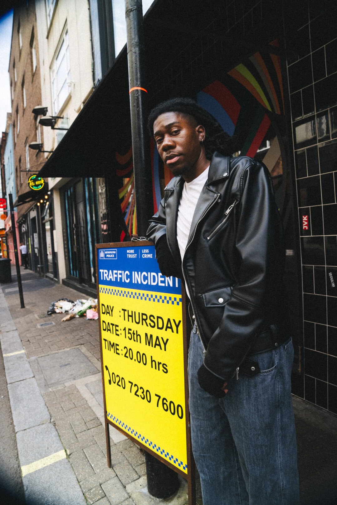Urban street photography featuring a young man in a black leather jacket near a traffic incident sign in a city environment.