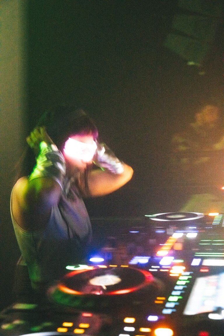 A DJ with a light-up face mask performing in a club with colourful lighting effects.