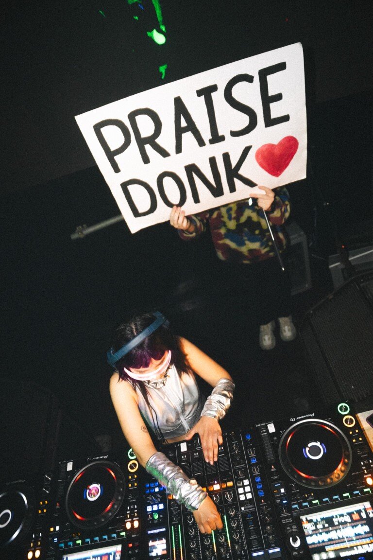 Pledge applause sign with a red heart held by a person wearing patterned clothing and playing DJ equipment, encouraging positivity and celebration at a musical event.