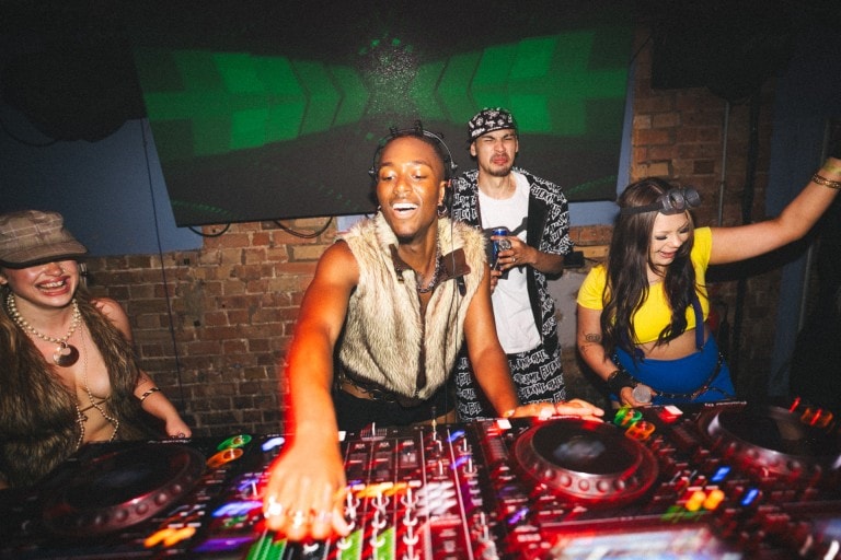 Vibrant club scene showing diverse partygoers dancing and DJing with colourful lighting, lively atmosphere, and a textured brick wall background.