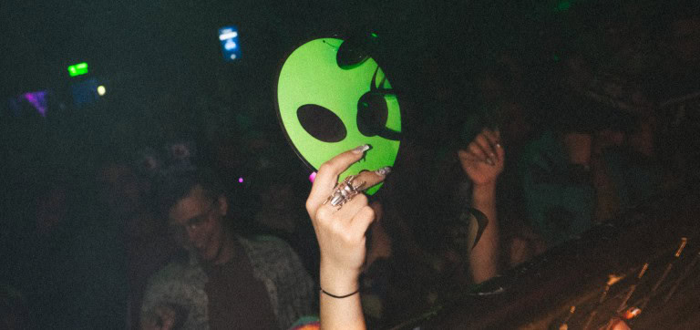 Alien face mask in neon green held by a person with silver rings at a dark party scene with dancing individuals in the background.