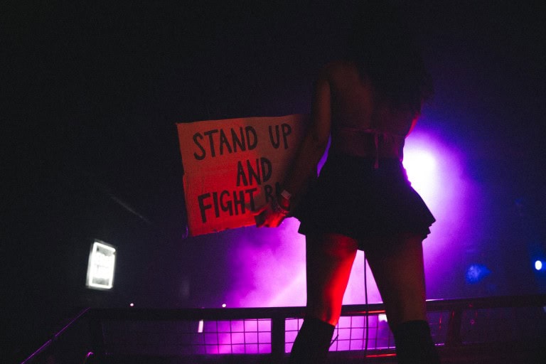 Person holding a protest sign with "STAND UP AND FIGHT" written on it, illuminated by colourful stage lights, capturing a moment of activism and empowerment.