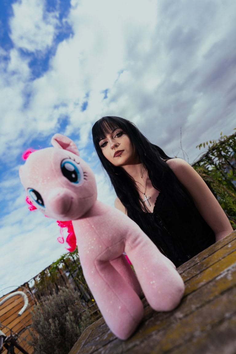 Person with long dark hair and edgy fashion holding a giant plush unicorn, set outdoors under stormy clouds.