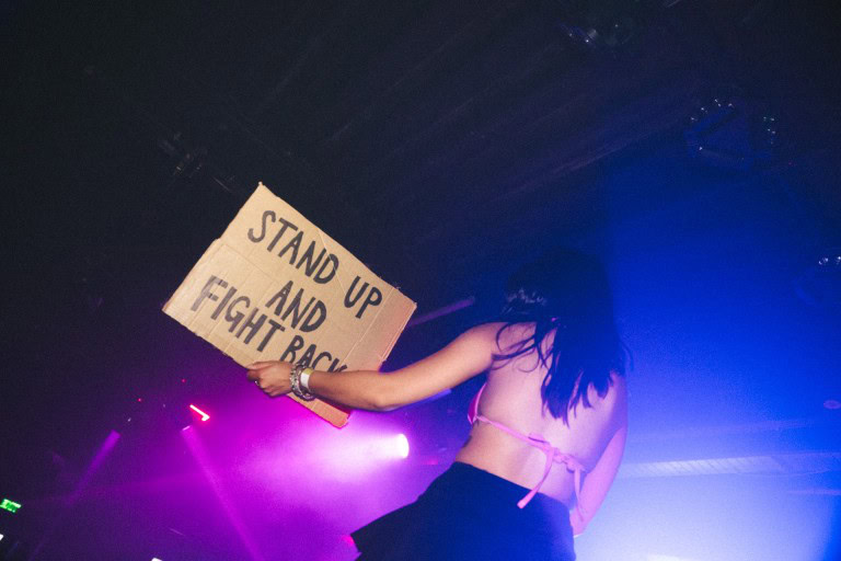 Protestor holding a handmade sign reading "Stand Up and Fight Back" during a rally under colourful stage lighting.