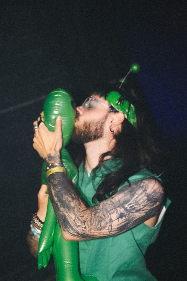 Balloon bouquet held by a person with tattoos, kissing the top of a green balloon, wearing protective glasses, with dark hair and a headband, in a lively environment.