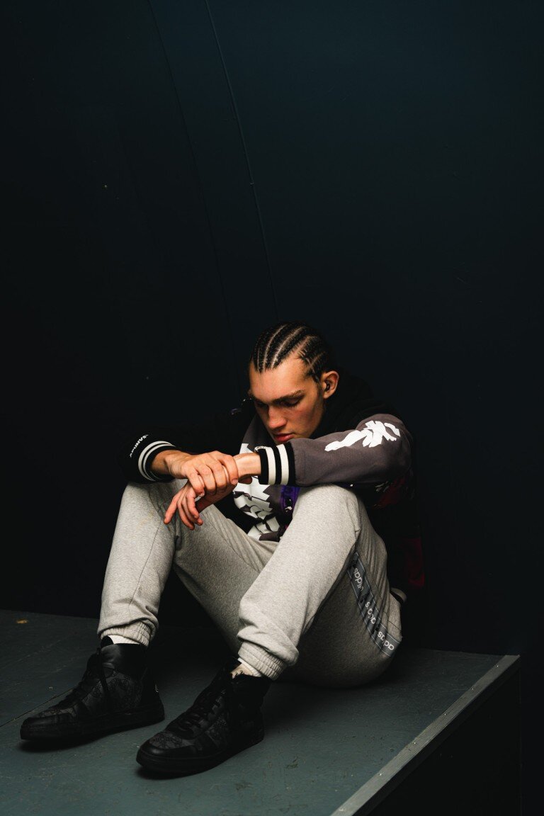 Wearing a black and grey hoodie with white graphic elements, grey joggers with branding details, and black high-top sneakers, a person sits on the floor with a dark background.