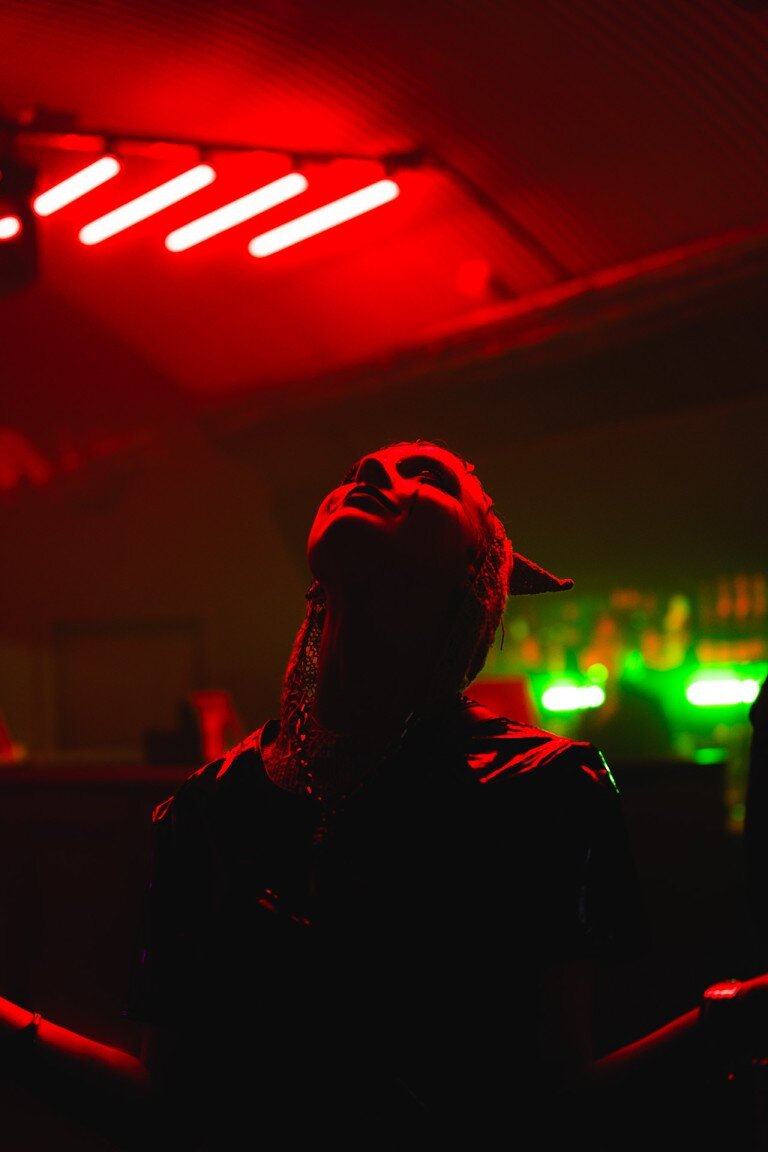 Vivid red lighting highlights an individual with dreadlocks and face paint in a darkened venue, creating an atmospheric and intense scene.