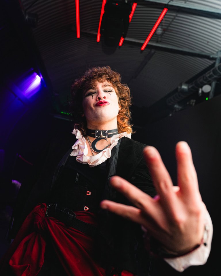 Expressive individual with bold makeup and alternative fashion, striking a confident pose in a vibrant nightclub setting.