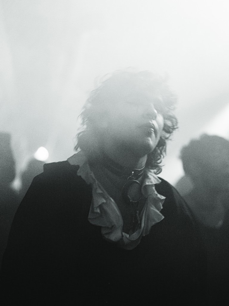 Mysterious person with wavy hair in black and white, wearing a ruffled shirt and headphones around neck, surrounded by a foggy atmosphere.