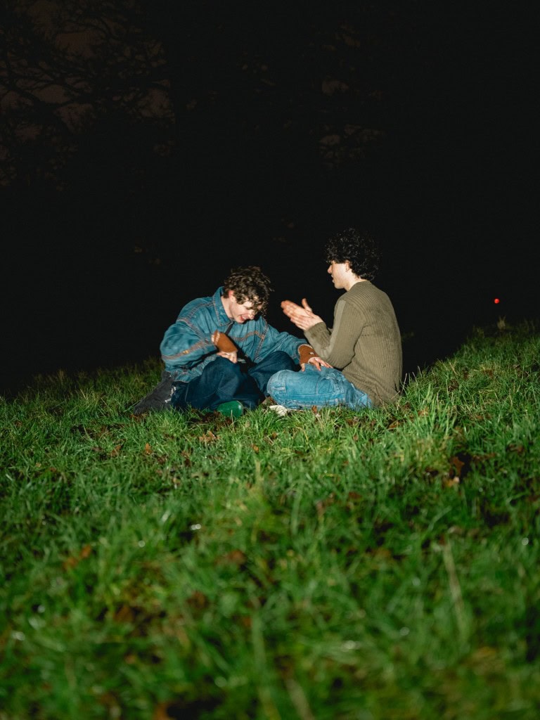 A young couple with curly hair sitting on a grassy hill at night, sharing a joyful moment and clapping hands, surrounded by dark trees.