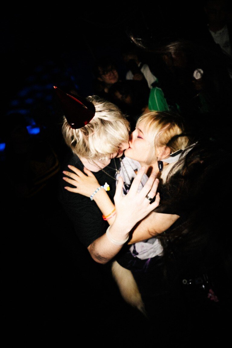 Kissing couple at a celebration event, capturing candid joy, group party, happy moments, night party, lively atmosphere, event photography, candid shot, emotion, young adults.
