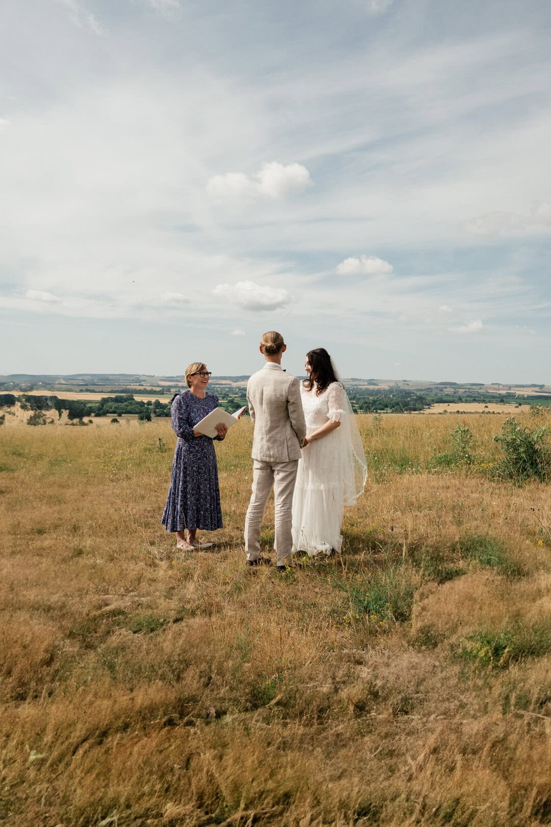 Beautiful outdoor wedding ceremony with bride, groom, and officiant in a scenic rural field under blue skies.