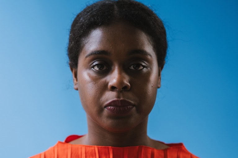 Close-up of a confident individual with short curly hair and deep brown skin, wearing a vibrant red top against a plain blue background.