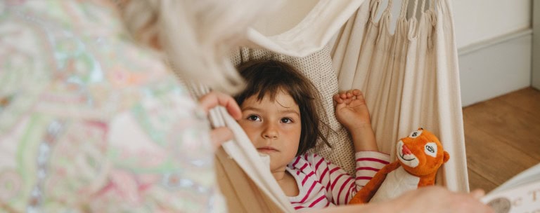 Young person with dark hair looking up from hammock with a plush tiger toy, indoor setting, relaxed and contemplative mood.