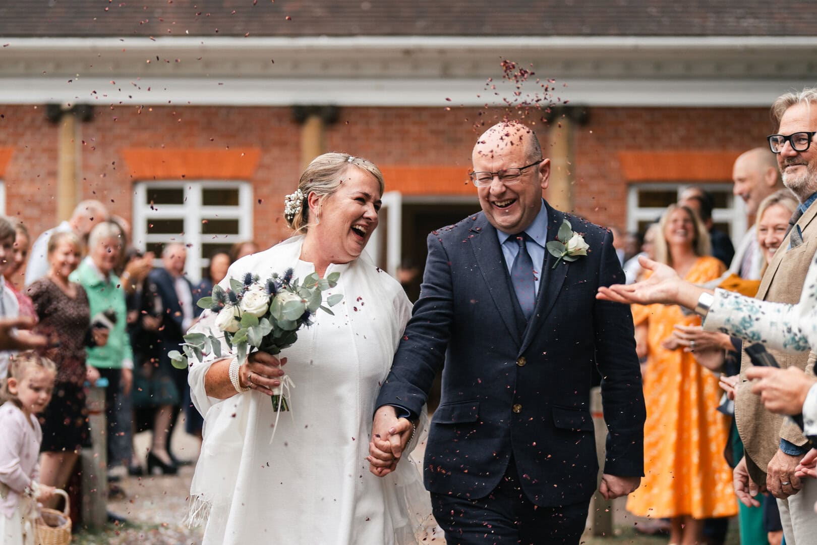 Brightly dressed people celebrating a wedding outdoors, with confetti and joyful expressions, in front of a rustic brick building.