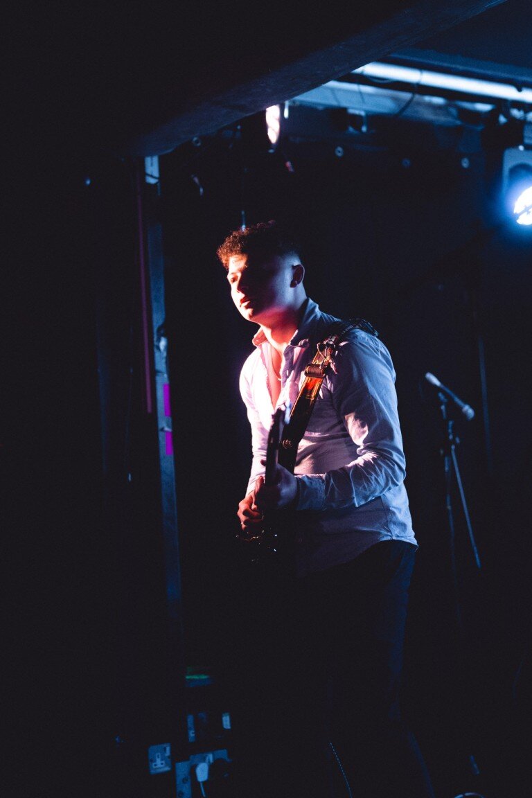 Guitarist performing on stage under dramatic lighting, dressed in a white shirt, creating a captivating atmosphere during the live music event.
