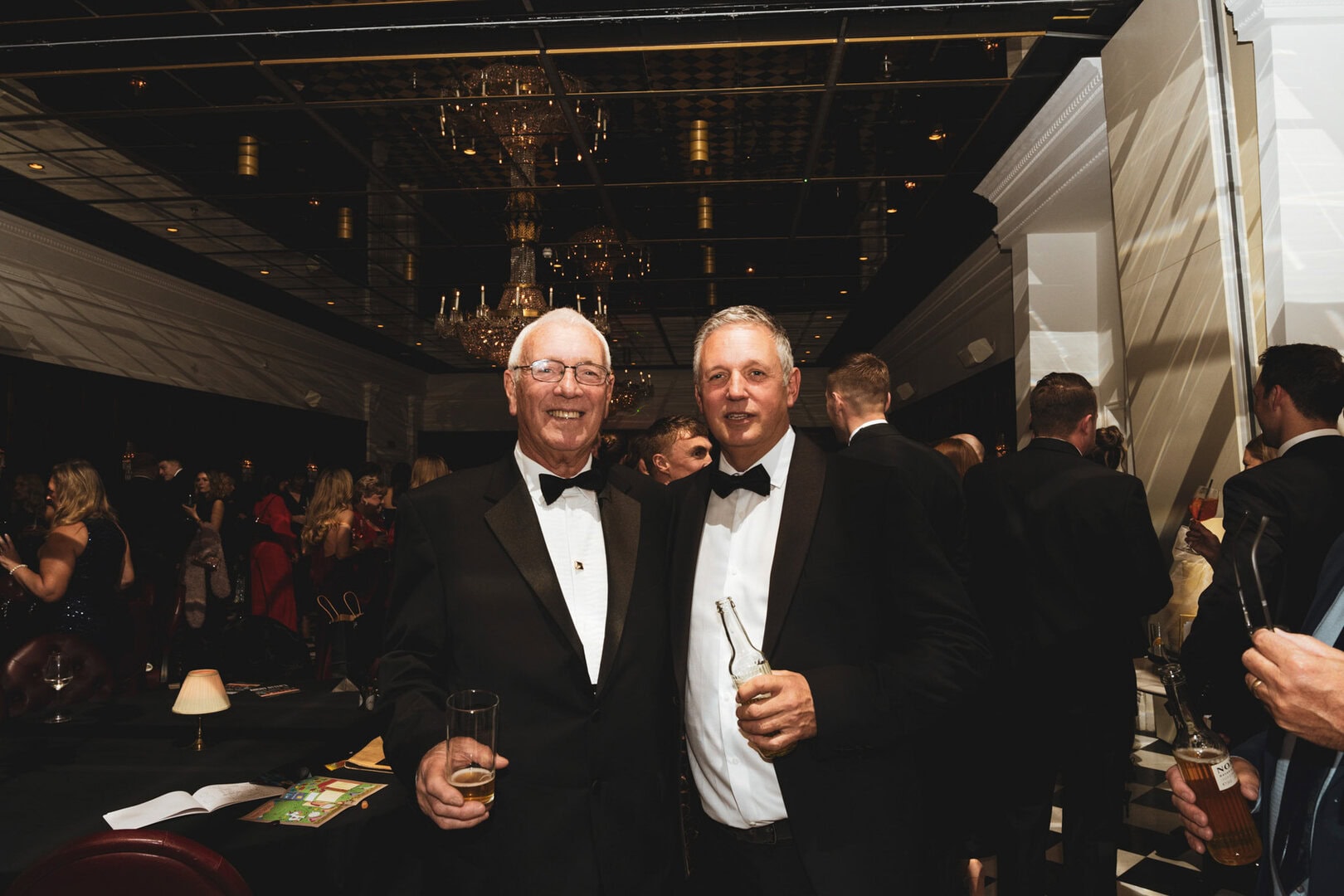 Elegant individuals in formal tuxedos at a black-tie event with sophisticated decor and chandeliers.